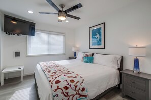 A bedroom with a bed, a ceiling fan, a TV mounted on the wall, a window with blinds, a bedside table, a nightstand with a lamp, and a framed picture on the wall.