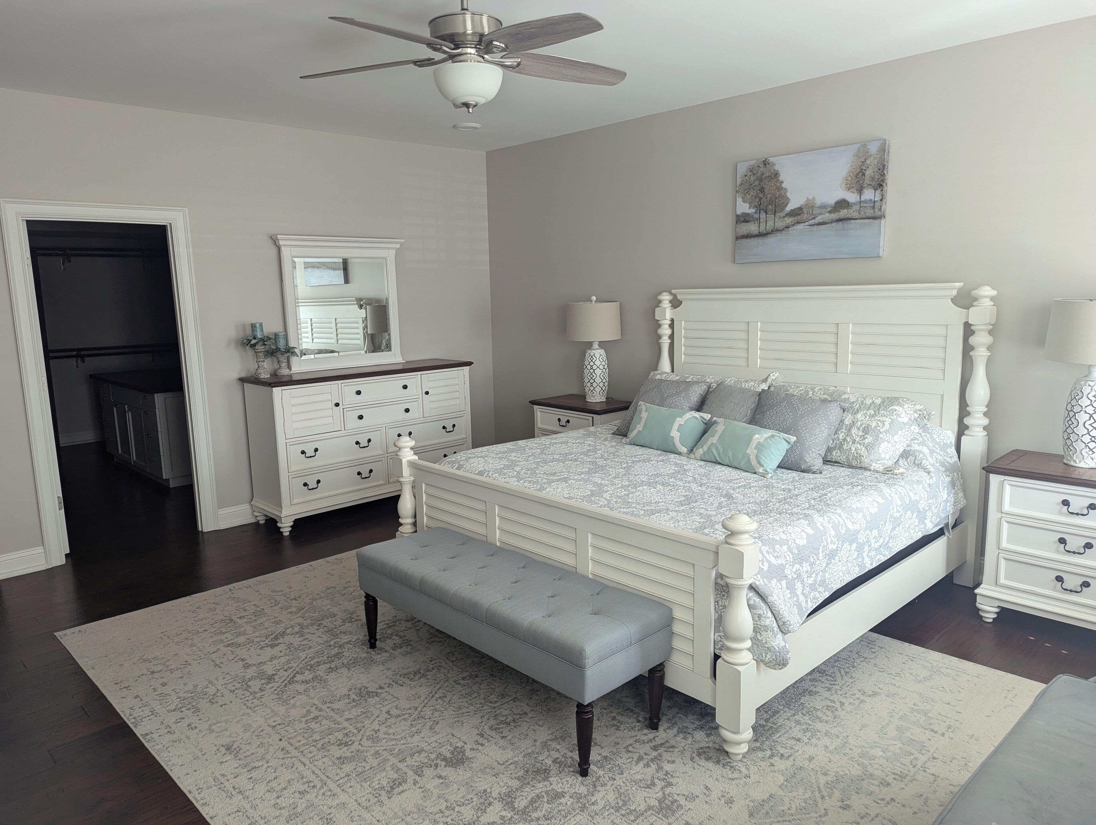 Une chambre à coucher comprenant un lit, une table de chevet, une commode, un miroir, un ventilateur de plafond et un tableau accroché au mur.
