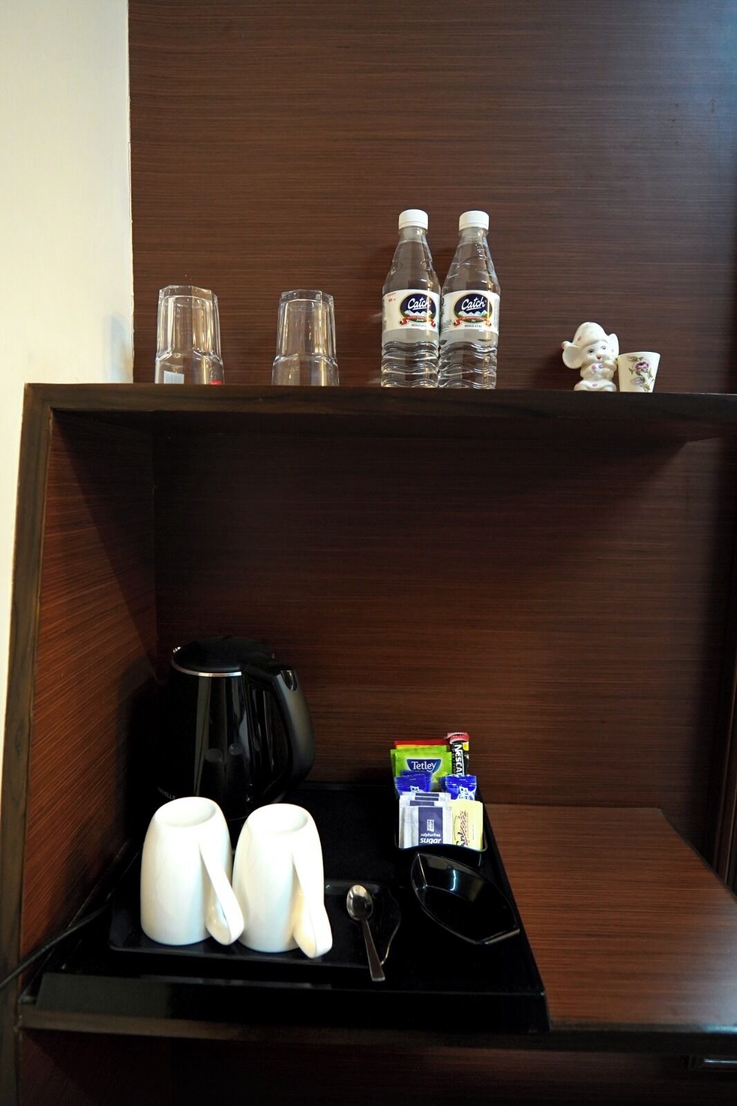 Quarto de hotel com uma prateleira de madeira contendo duas garrafas de água, dois copos e uma pequena estatueta. Abaixo, um serviço de mesa com uma cafeteira, um bule, xícaras, uma colher e um açucareiro.