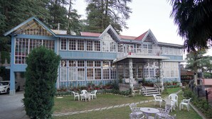 A large blue building with a balcony, surrounded by a garden with white chairs and tables. - The Edgeworth (Shimla)