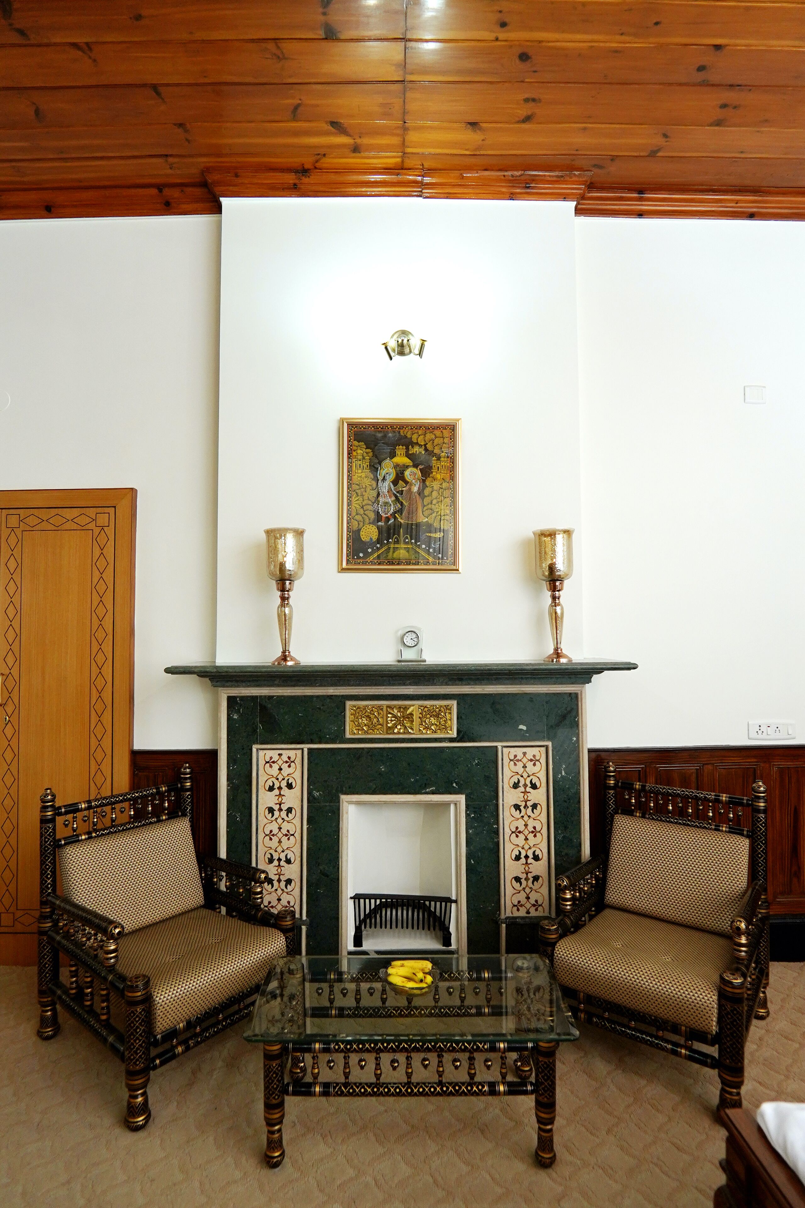 A room with a fireplace, two ornate chairs, a glass coffee table, and a framed painting above the mantel.