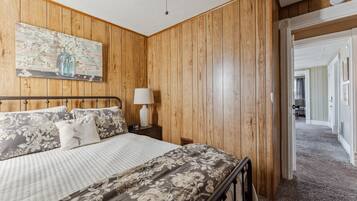 A bedroom with wooden paneling, a bed with floral bedding, a nightstand with a lamp, and a painting on the wall.