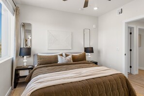 Une chambre à coucher avec un grand lit, deux tables de chevet avec des lampes, un miroir fixé au mur et une porte donnant sur une autre pièce.