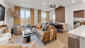Ein großzügiger Wohnbereich mit Sofa, Couchtisch und Esstisch. Ein großes Fenster mit Vorhängen, ein Deckenventilator und ein Blick ins Freie.
