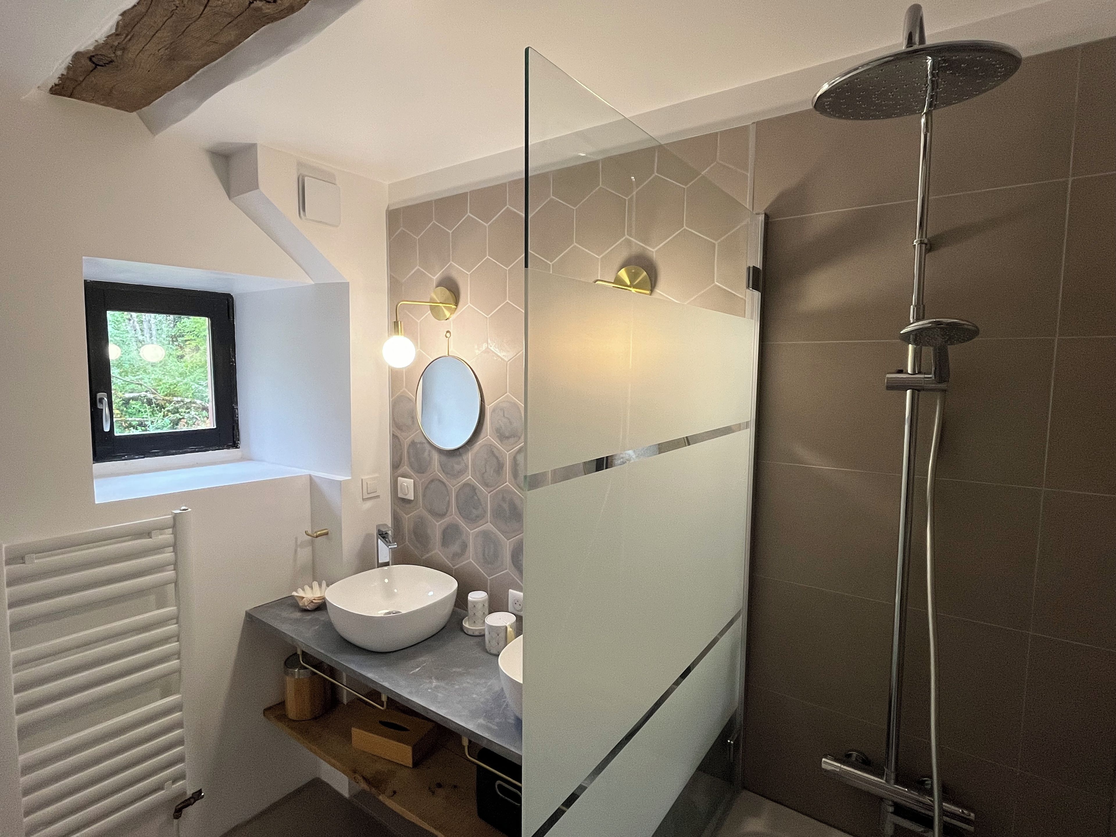 Un baño con un lavamanos blanco, un espejo redondo, una ducha con puerta de cristal y una ventana con vistas a la vegetación.
