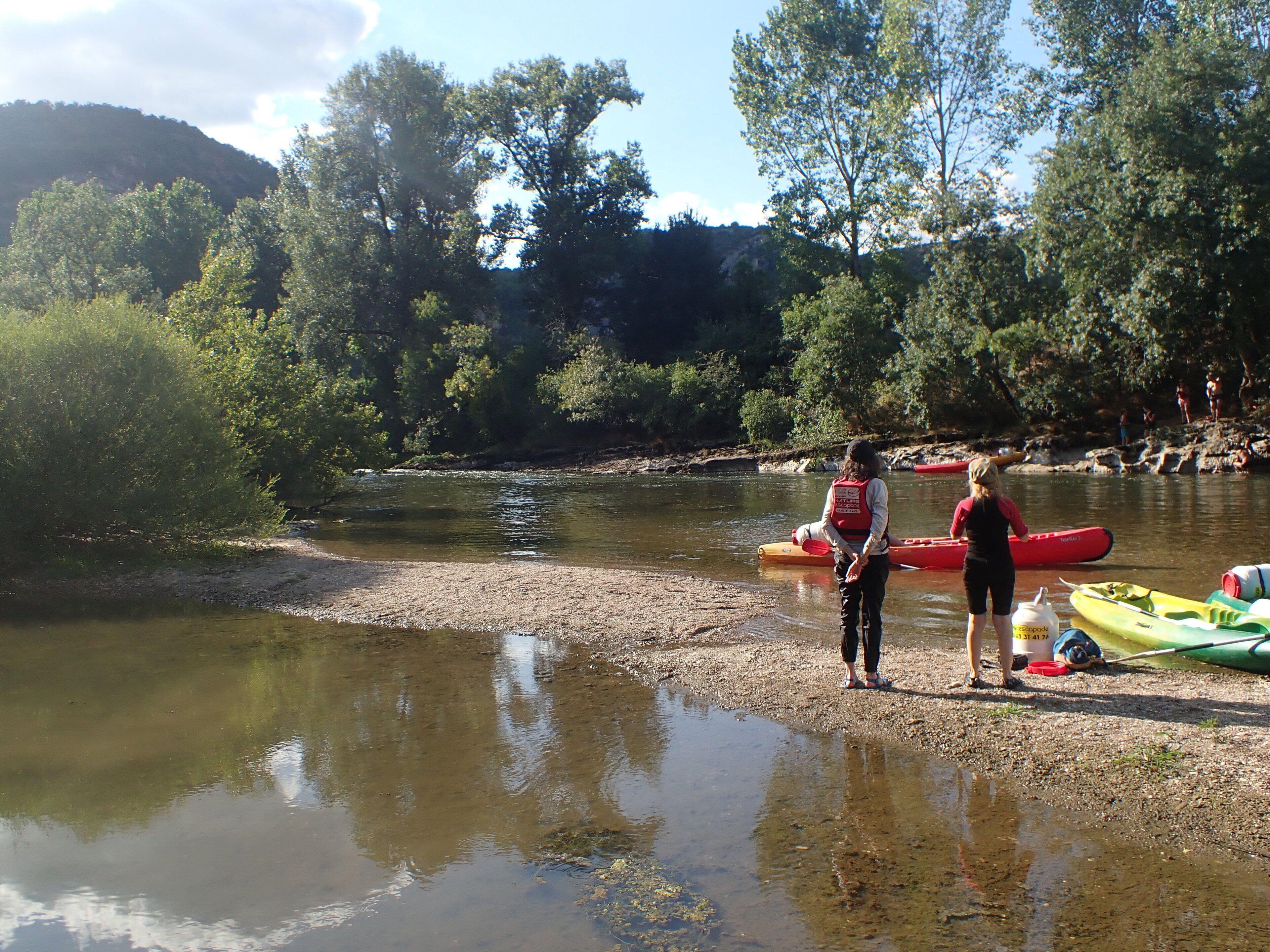 Orilla de río con kayaks y una persona con chaleco salvavidas.