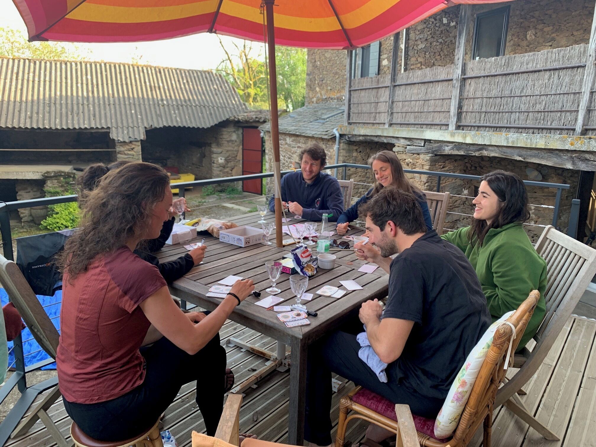 Un groupe de personnes est assis autour d’une table en bois à l’extérieur, avec un bâtiment en pierre en arrière-plan.