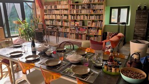 Un comedor con una mesa dispuesta para dos personas, rodeada de sillas, una estantería y un ventanal con cortinas.