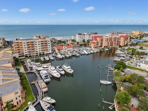Marina con varias embarcaciones amarradas, rodeada de edificios residenciales y una playa al fondo.