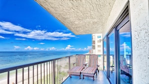 A balcony with a view of the ocean, two striped lounge chairs, and a glass door leading to a pool area. - 🌞Beachfront🌊Ocean Sands I-808🌴Private Balcony🏖️Amazing Beach Views (Madeira Beach)