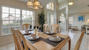 A dining area with a wooden table set for six, surrounded by chairs. There are large windows with blinds, a potted plant, and a chandelier above the table. - Spacious 4-bed lakeside pool villa near Disney (Kissimmee)