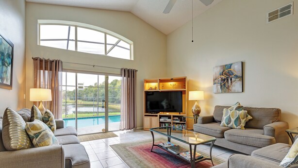 A spacious living room with a large TV, comfortable seating, and a view of a pool. - Spacious 4-bed lakeside pool villa near Disney (Kissimmee)
