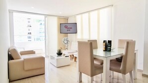 A modern living room with a glass dining table, beige sofa, and a flat-screen TV mounted on the wall. - PARCEROS GROUP - 2 BEDROOM VACATION APARTMENT, 10TH FLOOR, CARTAGENA, SPAIN (Cartagena de Indias)