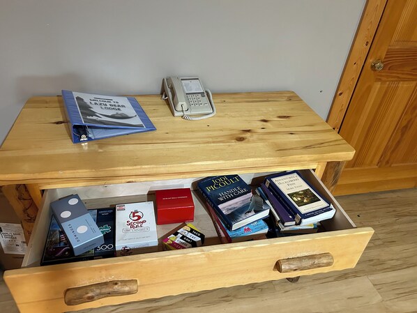 Ein Holztisch mit Telefon und Broschüre darauf. Darunter befindet sich eine Schublade mit verschiedenen Gegenständen, darunter Bücher, eine rote Schachtel und eine Schachtel mit der Aufschrift '5-Sekunden-Regel'.