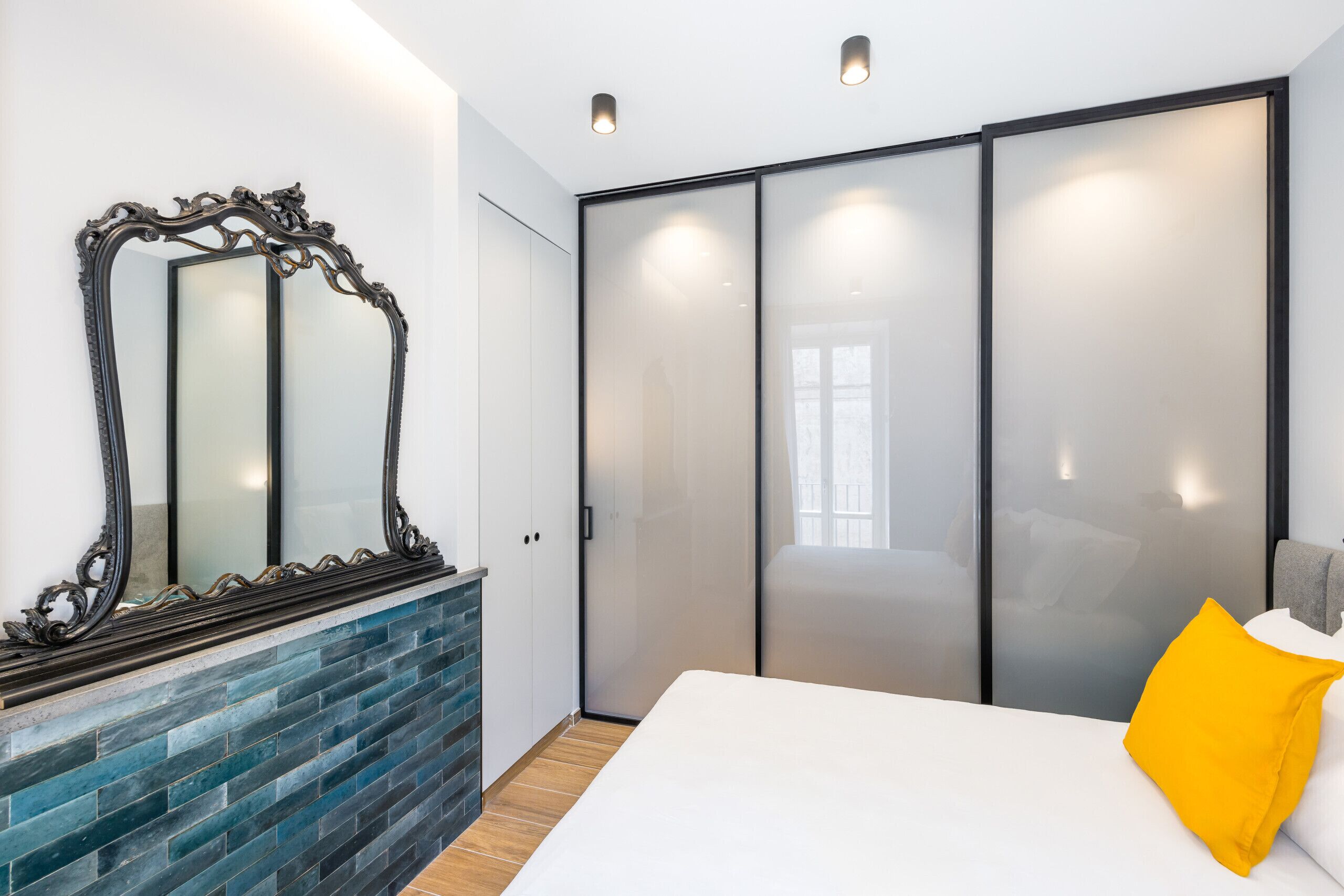 A bedroom with a white bed, a large mirror, a blue-tiled fireplace, and a sliding glass door closet.