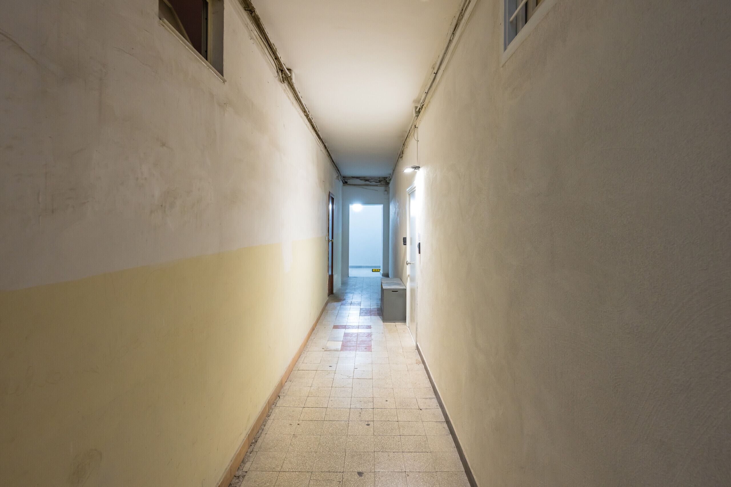 En smal hall med enkla väggar och ett kaklat golv, som leder fram till en dörr i slutet.