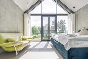 A modern bedroom with a large sliding glass door to a balcony, a green sofa, and a bed with a blue headboard. - Ferienhaus in Fuhlendorf am Boddenhafen (Fuhlendorf)