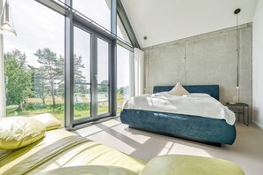 A modern bedroom with a large bed, a view of greenery outside, and a concrete accent wall. - Ferienhaus in Fuhlendorf am Boddenhafen (Fuhlendorf)