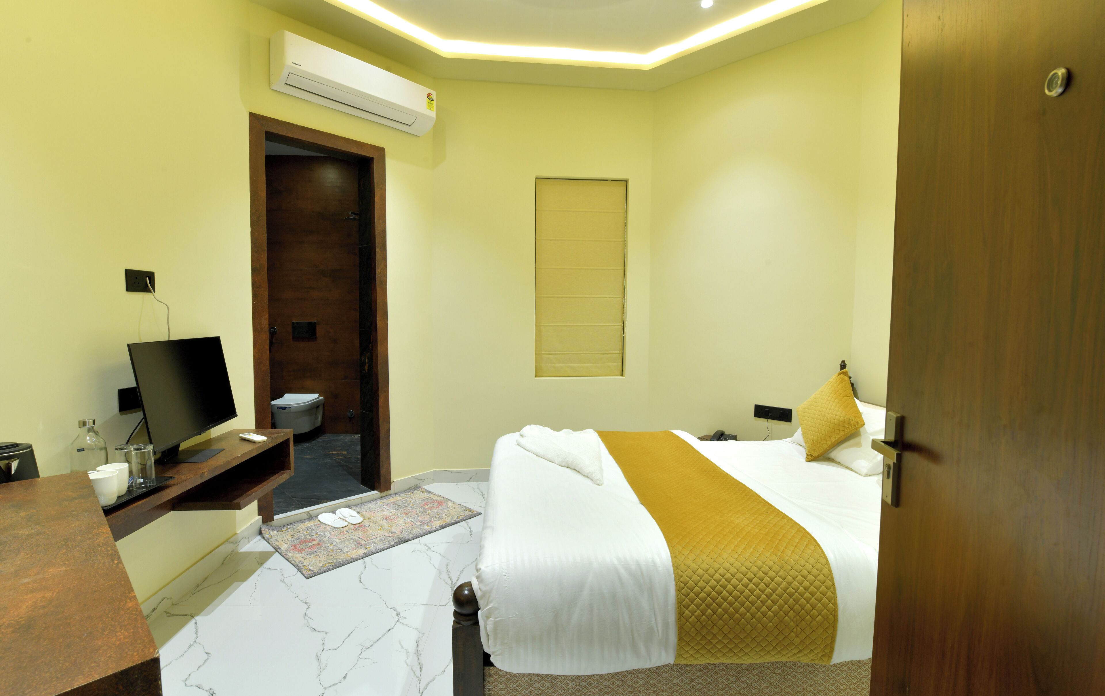 A hotel room with a bed, bedside table, TV, and an air conditioning unit.