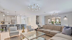 A modern living room with a sofa, armchairs, a glass coffee table, and a kitchen area in the background. - 9BR house with private pool in Champions Gate (Kissimmee)