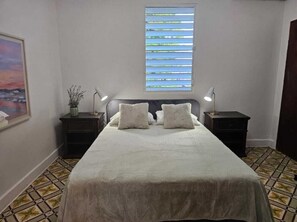 A bedroom with a bed, two bedside tables, a nightstand, a window with blinds, and a framed picture on the wall.