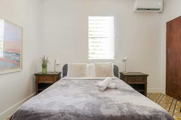 A bedroom with a bed, two bedside tables, a nightstand, a window, and an air conditioning unit.