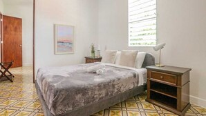 Un dormitorio con cama, mesita de noche, ventana con persianas, un cuadro enmarcado en la pared y una puerta.