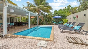 Une piscine avec une terrasse carrelée, des chaises longues dotées de coussins à motifs, un espace salon couvert et un hamac.
