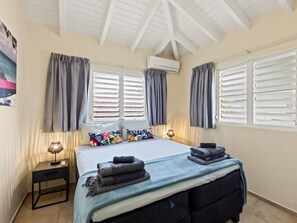 A bedroom with a bed, two bedside tables, an air conditioning unit, and a window with shutters.