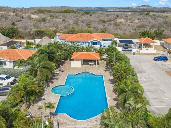A resort with a central swimming pool, multiple villas with red-tiled roofs, and a parking area.
