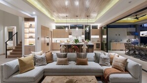A modern living room with a grey sectional sofa, a coffee table, and a kitchen in the background. - Mauna Lani Luxe Estate at One Ocean by Gather w/ Infinity Pool & Spa (Waimea)