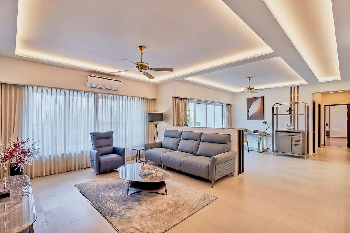 Una sala moderna con un sofá gris, un sillón negro y una mesa de centro. Se ven ventiladores de techo, un televisor y un comedor.