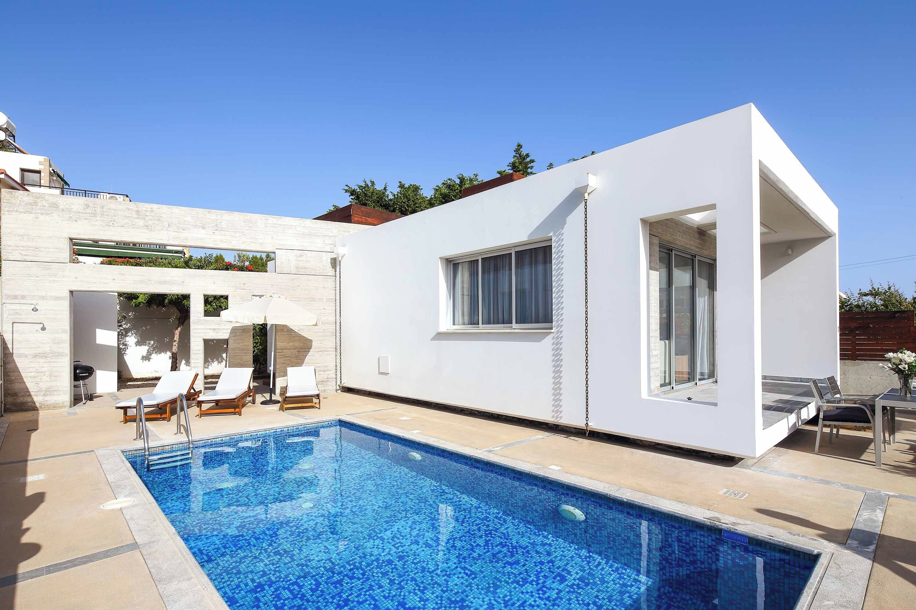 Uma casa moderna com piscina, fachada branca e um terraço com cadeiras de descanso.