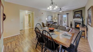 A spacious dining area with a wooden table, black chairs, and a chandelier. There is a fireplace with a TV on a stand, and a view of a balcony with outdoor furniture. - Spacious 3-BR Condo for 8 w Resort Pools, Hot Tub, and Free Dollywood Ticket (Pigeon Forge)