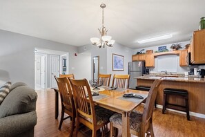 Un comedor con una mesa y sillas de madera, una lámpara de araña y una cocina al fondo.