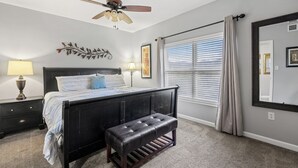 Une chambre à coucher comprenant un lit, un banc, un ventilateur de plafond, une fenêtre avec des stores et des éléments de décoration murale.