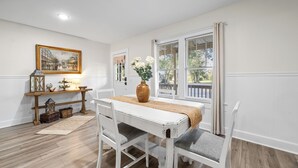 Un coin repas comprenant une table blanche, des chaises grises à coussins, une console en bois agrémentée d’objets décoratifs et une vue par une fenêtre équipée de stores.