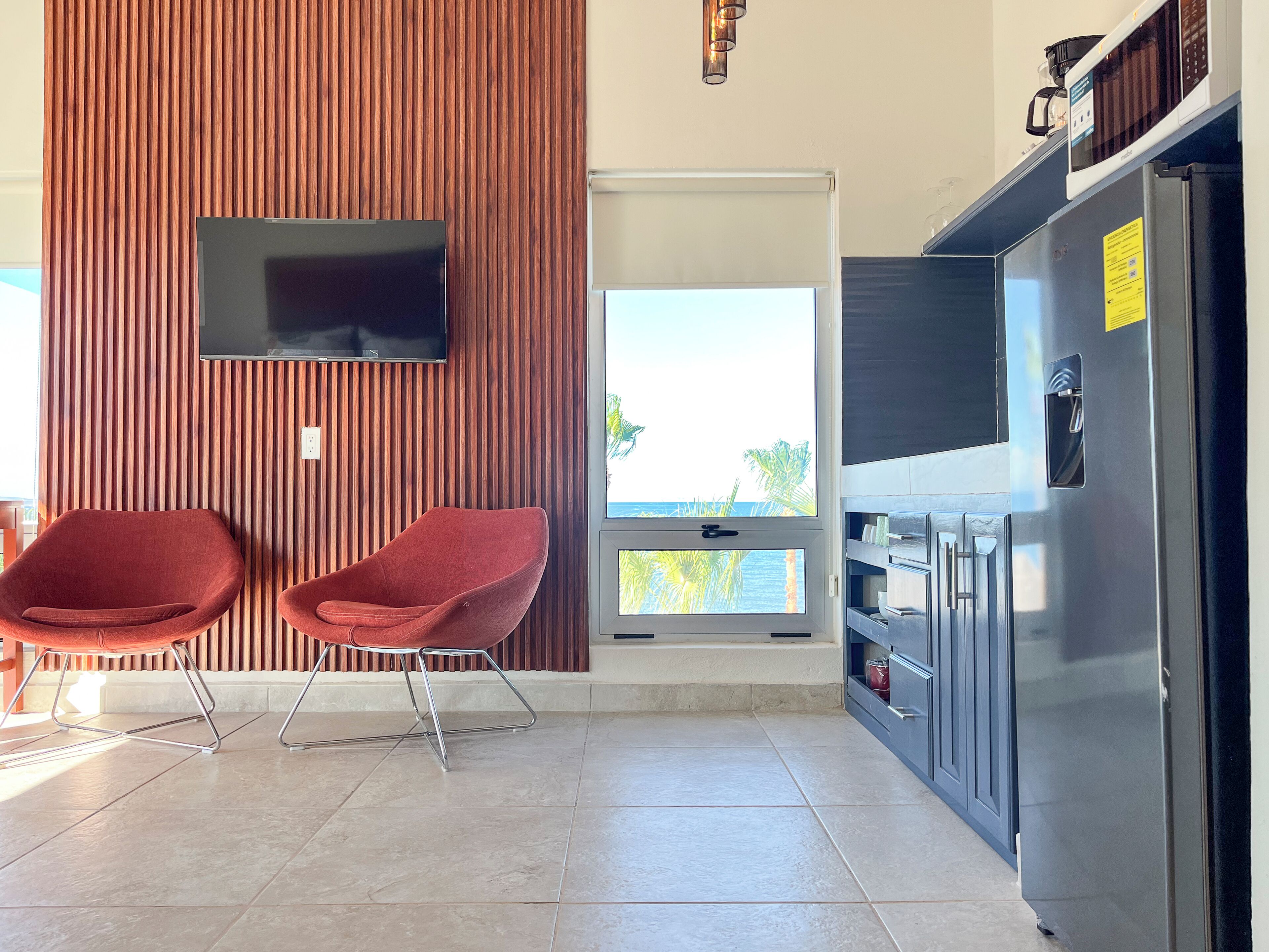 A modern kitchen with a built-in TV, two red chairs, a large window with a view, and a stainless steel refrigerator.