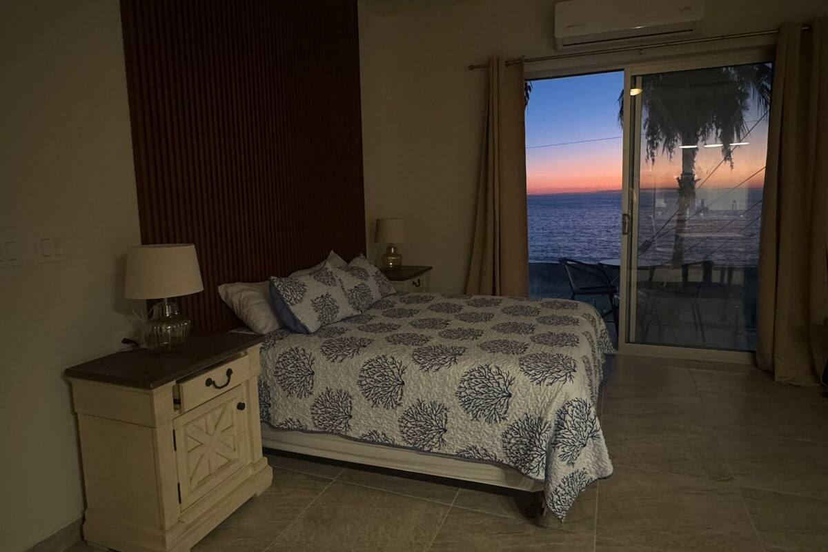 Un dormitorio con cama, mesita de noche, lámpara y vista al océano al atardecer.