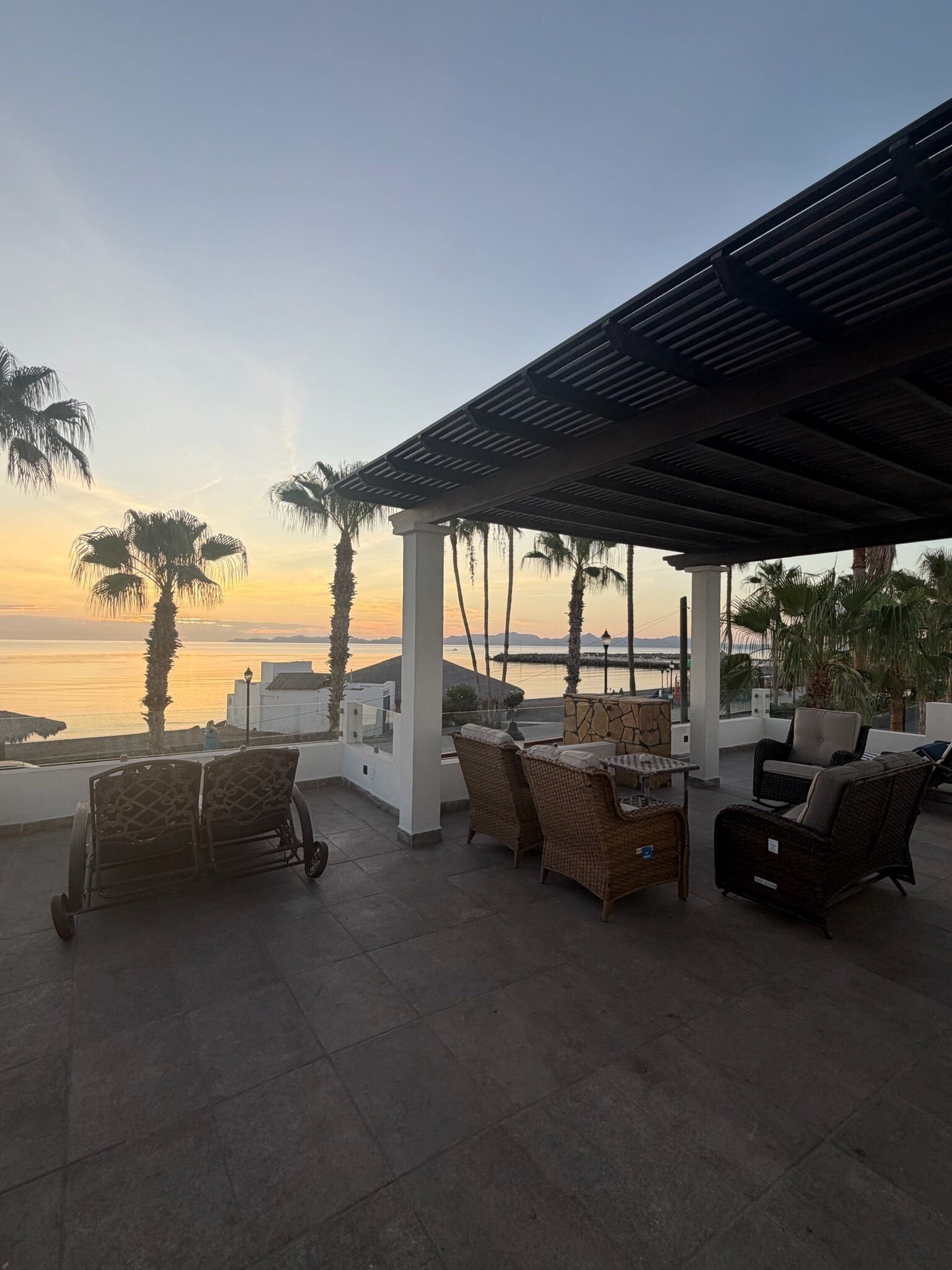 A covered outdoor seating area with wicker furniture, a view of the sea, and palm trees.