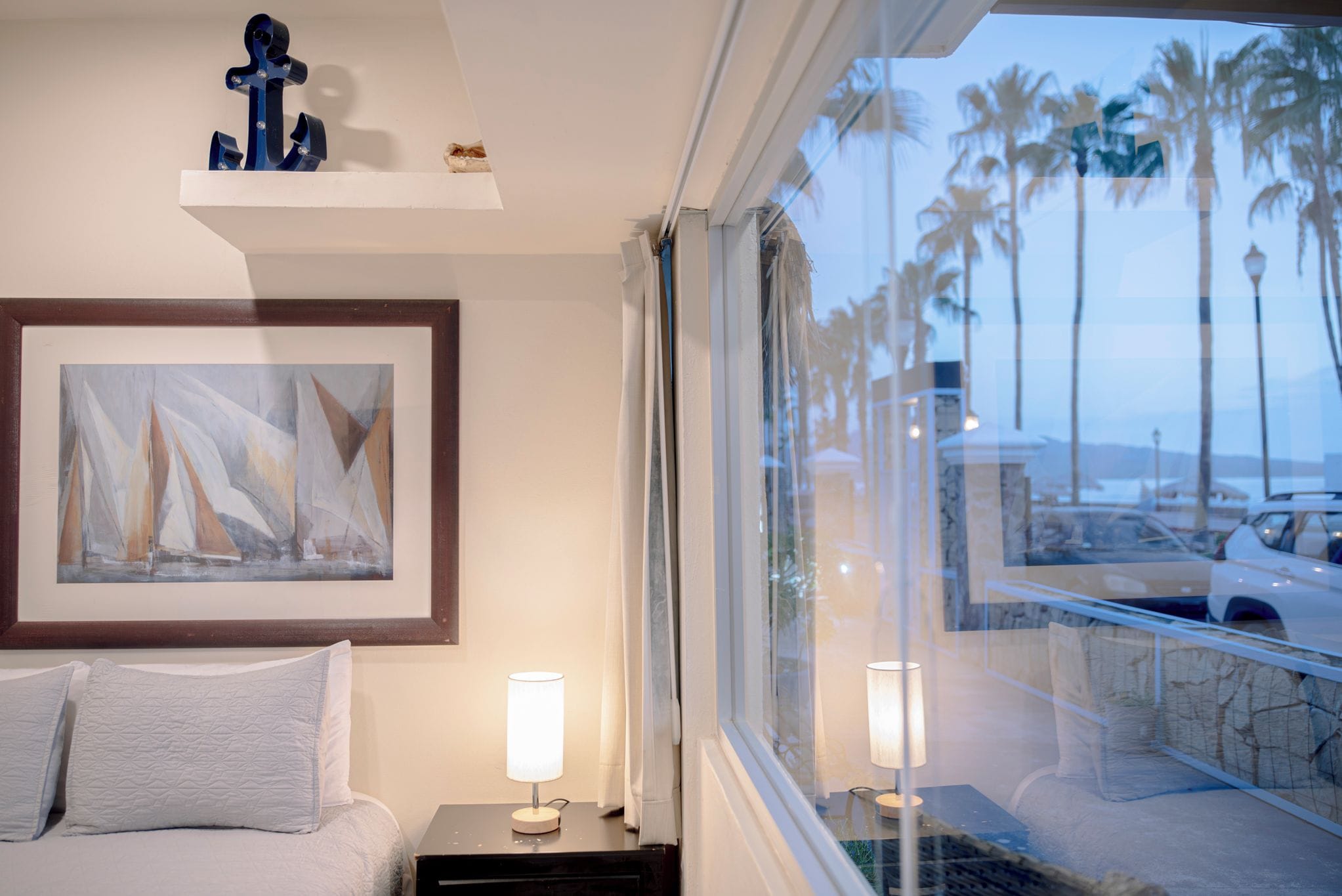 A hotel room with a bed, a framed abstract painting, a nightstand with a lamp, and a view of palm trees and a parking area through the window.