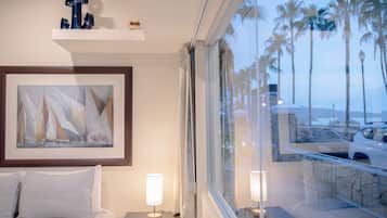 A hotel room with a bed, a framed abstract painting, a nightstand with a lamp, and a view of palm trees and a parking area through the window.