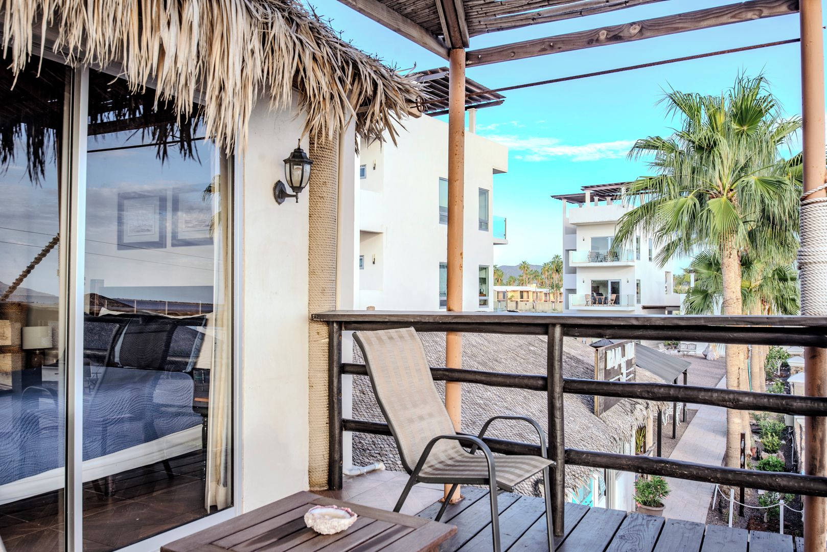 A balcony with a thatched roof, a table with a shell decoration, and a view of a coastal area with palm trees and buildings.