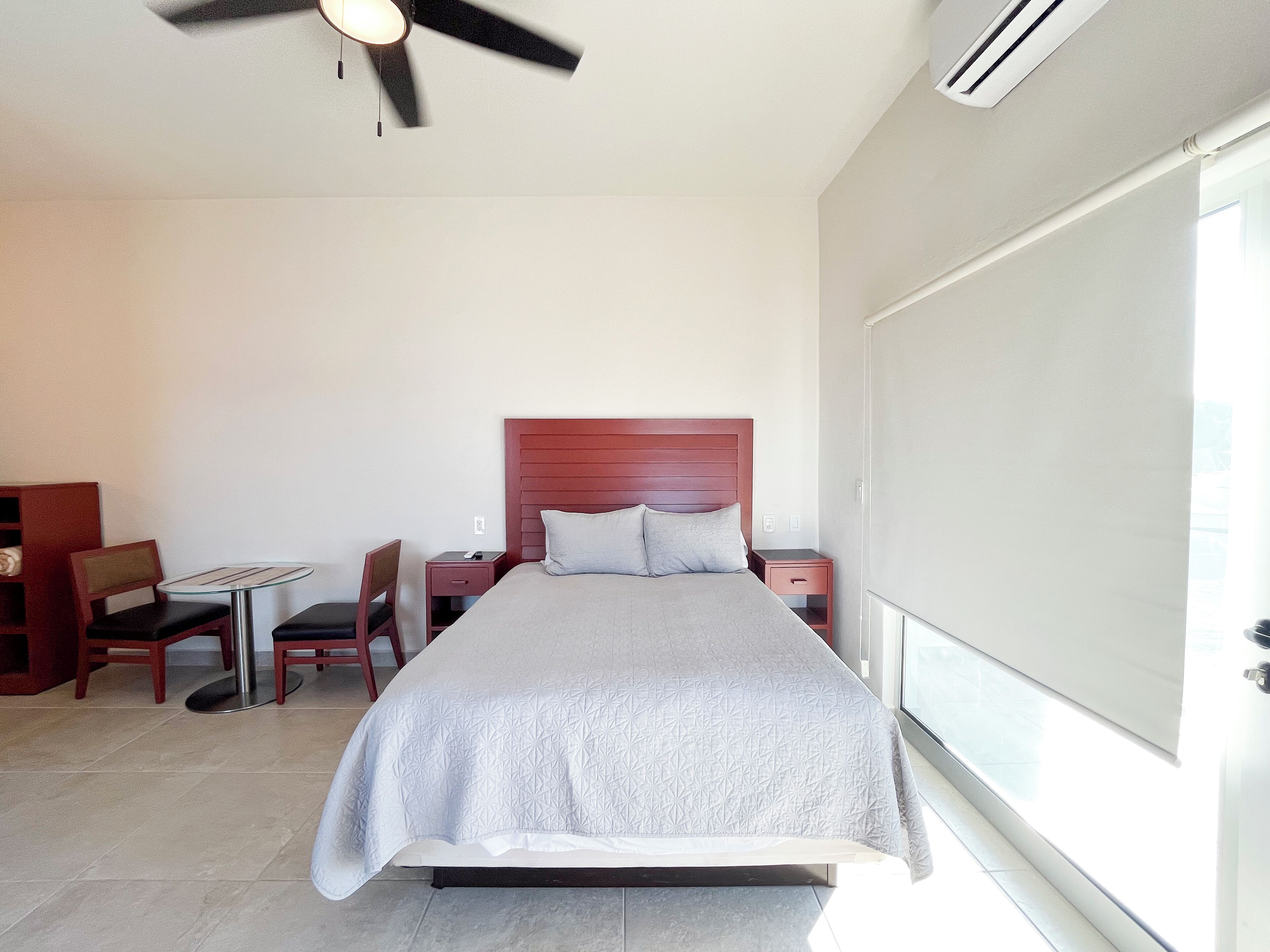 A hotel room with a bed, a desk, a chair, a small table, a ceiling fan, and a window.