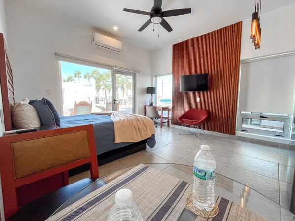 A hotel room with a bed, a desk, a chair, a TV, and a window with a view of palm trees. - BAJA MIA SUITES AND TOURS (Loreto)
