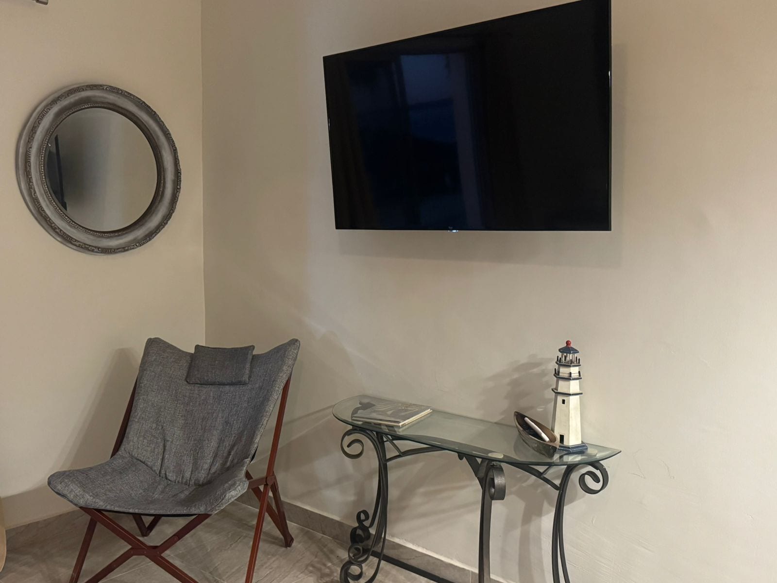 Une pièce comprenant un fauteuil recouvert d’un tissu gris, un miroir rond, une table en verre ornée d’un objet décoratif et un téléviseur à écran plat fixé au mur.