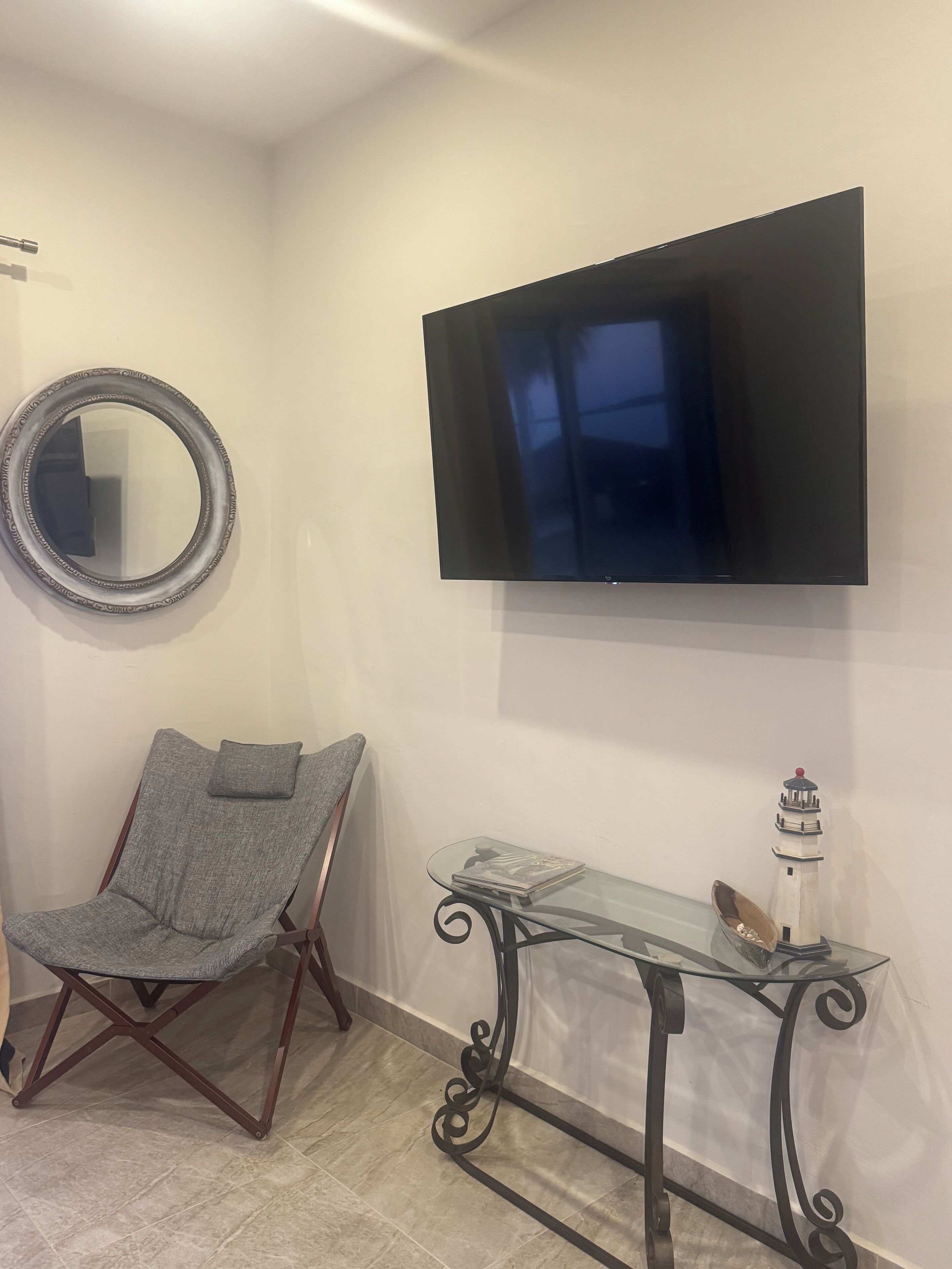Ein Wohnzimmer mit einem an der Wand befestigten Flachbildfernseher, einem runden Spiegel, einem grau gepolsterten Sessel und einem Glastisch mit einer Modellleuchtturms.