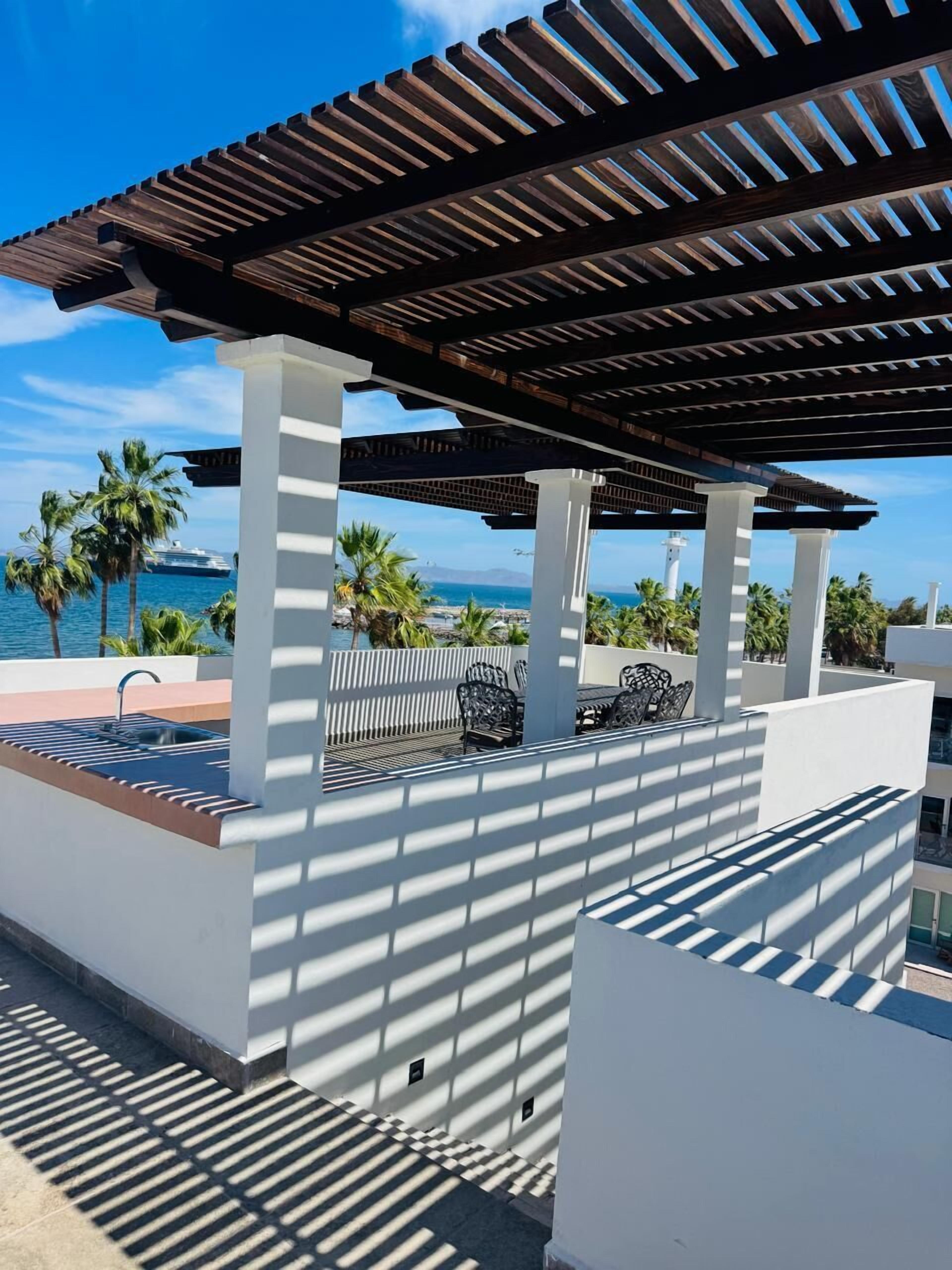 Un espacio al aire libre cubierto con zona de estar, piscina, columnas blancas y vistas al mar.