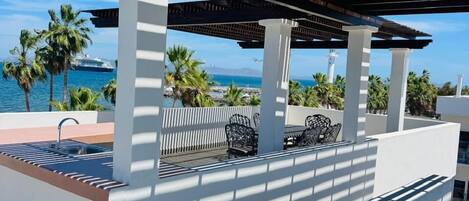 Un espacio al aire libre cubierto con zona de estar, piscina, columnas blancas y vistas al mar.