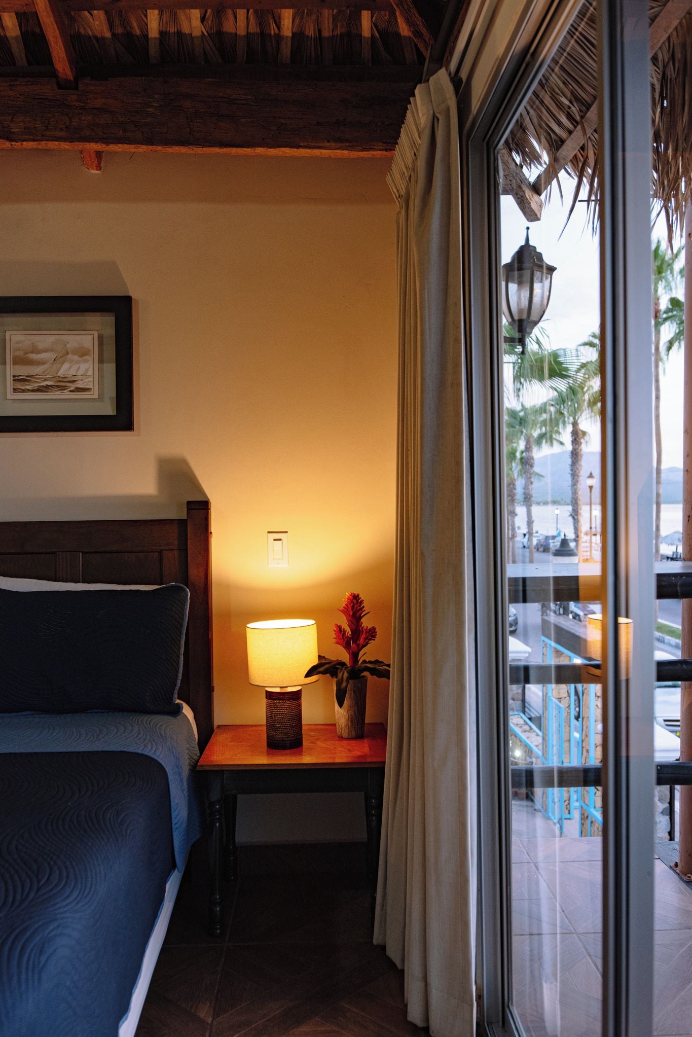 A hotel room with a bed, a nightstand with a lamp, a framed picture, and a view of the outside.